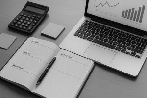 Black and white image of laptop, planner, and calculator on a desk for organized financial planning.